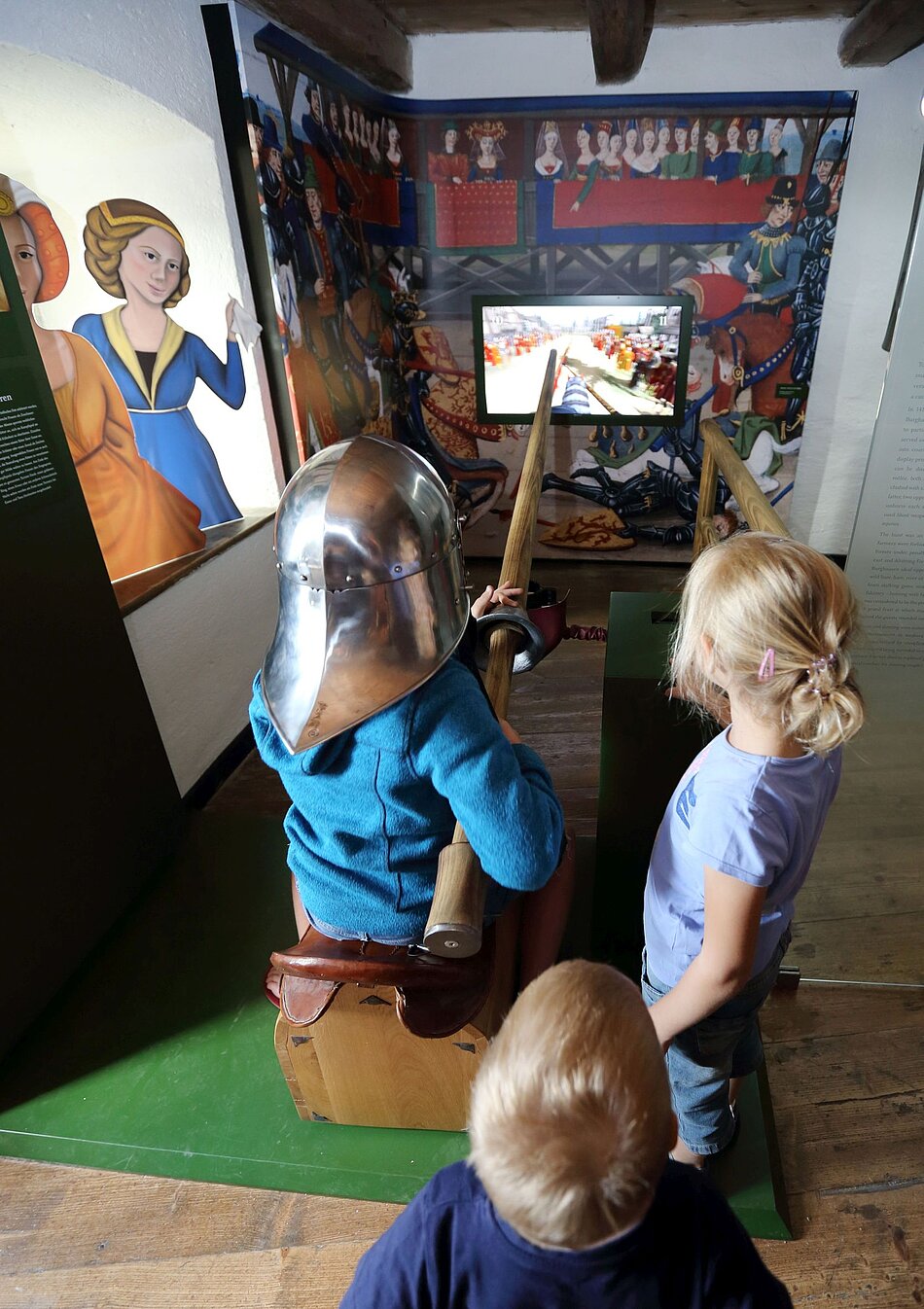Mehrere Kinder stehen vor einem Bildschirm. Eines der Kinder trägt eine Lanze und Ritterhelm und spielt ein Rittertunier nach.